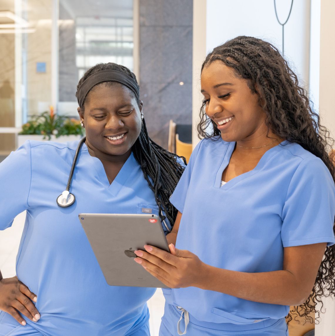 two female healthcare workers in scrubs smiling and using a tablet